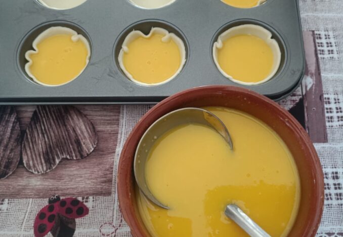 A close-up high-angle shot of freshly baked Pastel de Nata tarts on a rustic wooden table, featuring golden puff pastry crusts and caramelized creamy custard centers, dusted with cinnamon.