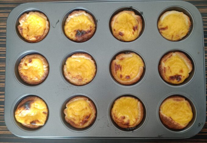 A close-up high-angle shot of freshly baked Pastel de Nata tarts on a rustic wooden table, featuring golden puff pastry crusts and caramelized creamy custard centers, dusted with cinnamon.